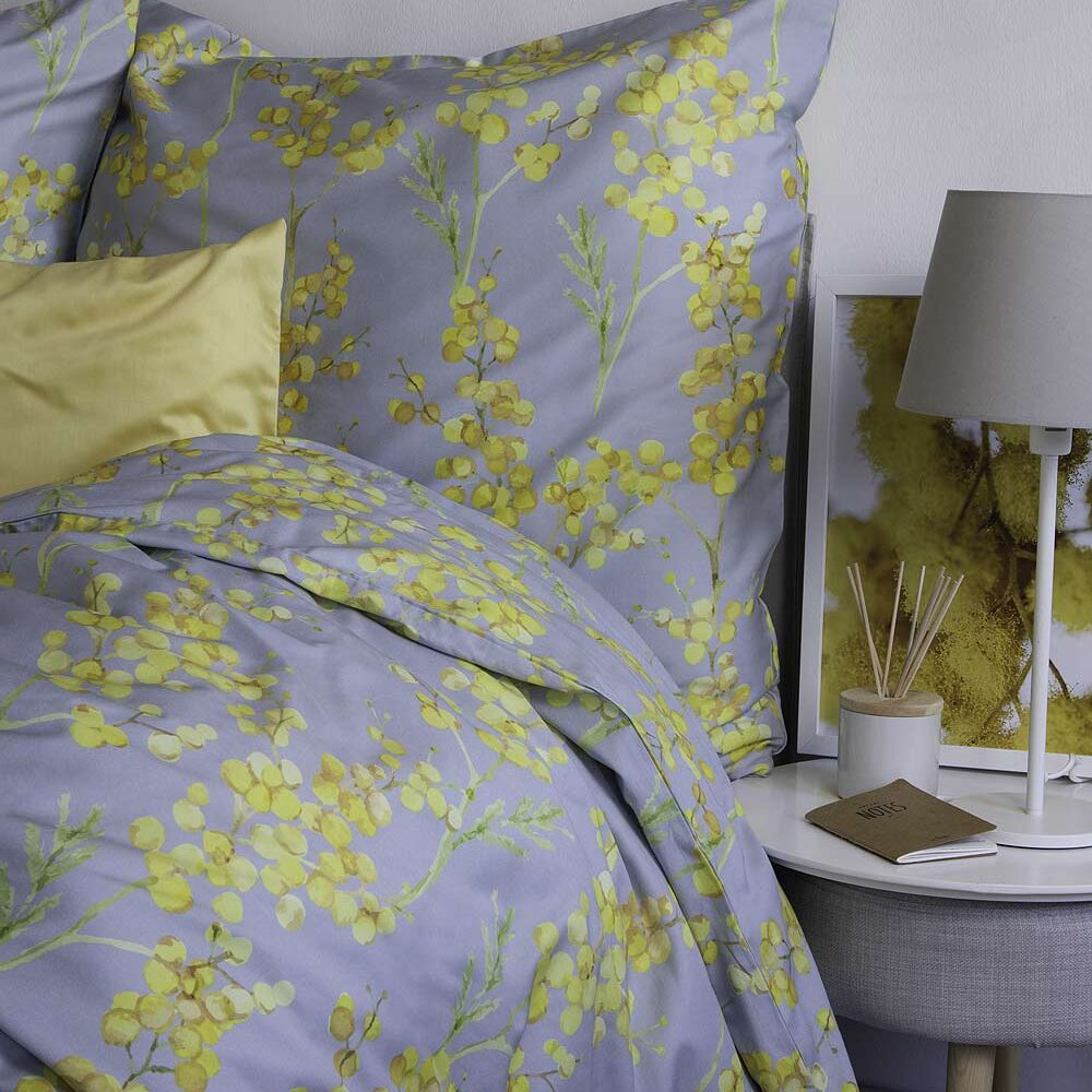 Bettwäsche Blüten fleuresse Ein gemütlich eingerichtetes Bett mit Bettwäsche in einem floralen Muster aus gelben Blüten und grünen Zweigen. Im Vordergrund sind ein großes Kissen und eine Decke zu sehen, beide mit dem gleichen floralen Muster. Daneben steht ein Nachttisch mit einer grauen Lampe, einem Duftstäbchen-Behälter, einem kleinen Buch mit dem Titel „Notes“ und einem gerahmten Bild, das ebenfalls gelbe Blüten zeigt. Die Atmosphäre wirkt ruhig und einladend.