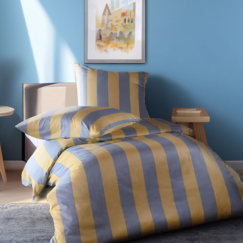 Modern Life Streifen Bettwäsche. Ein gemütlich eingerichtetes Schlafzimmer mit einem Bett, das mit gestreifter Bettwäsche in gelb und blau bezogen ist. Im Hintergrund ist eine hellblaue Wand mit einem gerahmten Bild, ein Nachttisch mit einem Buch und ein Teppichboden zu sehen. Natürliches Licht fällt durch ein Fenster ins Zimmer.