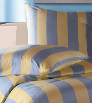 Modern Life Streifen Bettwäsche. Ein Bett, das mit gestreifter Bettwäsche in gelb und blau bezogen ist. Im Hintergrund ist eine hellblaue Wand. Die Bettwäsche wurde vom nahen fotografiert.
