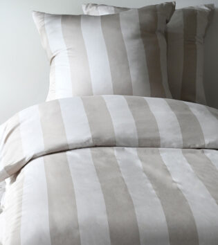 Modern Life Streifen Bettwäsche. Ein Bett, das mit gestreifter Bettwäsche in beige und weiß bezogen ist. Im Hintergrund ist eine hellblaue Wand. Die Bettwäsche wurde vom nahen fotografiert.
