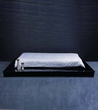 Das Bild zeigt ein Bett in einem dunklen Raum. Das Bett hat ein glänzendes, schwarzes, flaches Rahmen-Design, das fast wie ein Spiegel wirkt. Auf der Oberfläche liegt eine weiße Decke, die teilweise auf dem Bett hängt. Zwei schlanke, metallische Objekte stehen auf der Bettkante, die wahrscheinlich als dekorative Elemente dienen. Der Raum hat eine sehr schlichte, elegante Atmosphäre mit einem dunklen Hintergrund.