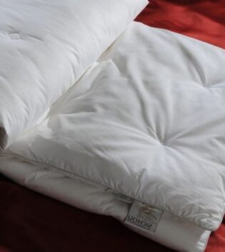 Das Bild zeigt eine weiße Decke oder Bettdecke, die locker auf einem roten Bett liegt. Es handelt sich um eine hochwertige, gefüllte Bettdecke mit gleichmäßigen, weichen Nähten, die die Füllung an Ort und Stelle halten. Das Material wirkt luxuriös und gut verarbeitet. Ein Etikett am Rand ist sichtbar, das die Marke oder den Hersteller zeigt.