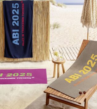 Drei Strandtücher mit dem Aufdruck „ABI 2025 – ALLES IN TROCKENEN TÜCHERN“ liegen oder hängen an einem Strand. Ein dunkelblaues Handtuch mit roter Schrift hängt an einem Sichtschutz aus Bambus. Ein magentafarbenes Handtuch mit grüner Schrift liegt im Sand. Ein braunes Handtuch mit gelber und oranger Schrift ist auf einer Holzliege ausgebreitet. Im Hintergrund befinden sich Strand, Meer und ein Sonnenschirm. Auf einem kleinen Holztisch stehen ein Krug mit Zitronenwasser und zwei Gläser.