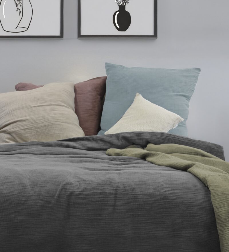 Ein dekoriertes Bett mit verschiedenen einfarbigen Bettwäschen und Kissen aus Musselin, lässig übergeworfen. Auf der Rechte Seite liegt ein kleines Kissen in creme. Im Hintergrund an der Wand hängen zwei passende Bilder.