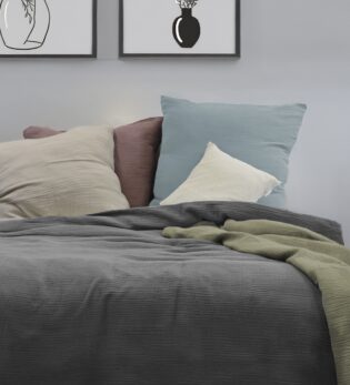 Ein dekoriertes Bett mit verschiedenen einfarbigen Bettwäschen und Kissen aus Musselin, lässig übergeworfen. Im Vordergrund liegt ein Bettbezug in salbei. Im Hintergrund an der Wand hängen zwei passende Bilder.