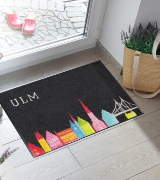 Wohnmatte mit schwarzem Hintergrund, graue Brücke und bunten Gebäuden in Blau, Gelb, Rot und Pink, Grün dazu das Ulmer Münster als zentrales Motiv.