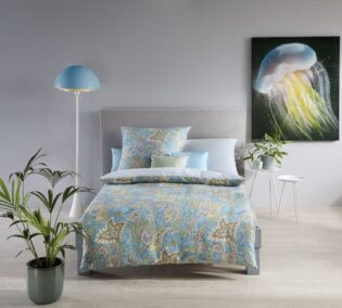 Modern eingerichtetes Schlafzimmer mit Bett in türkisfarbener Paisley-Bettwäsche. Neben dem Bett stehen eine blaue Stehlampe und eine Pflanze, an der Wand hängt ein Bild mit einer leuchtenden Qualle.